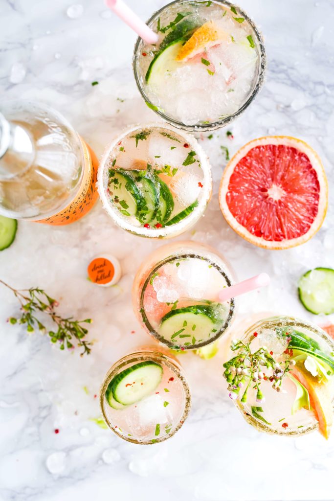 A variety of colorful cocktails displayed on a bed of ice with cucumbers , oranges, and sprigs of green as garnishes. 