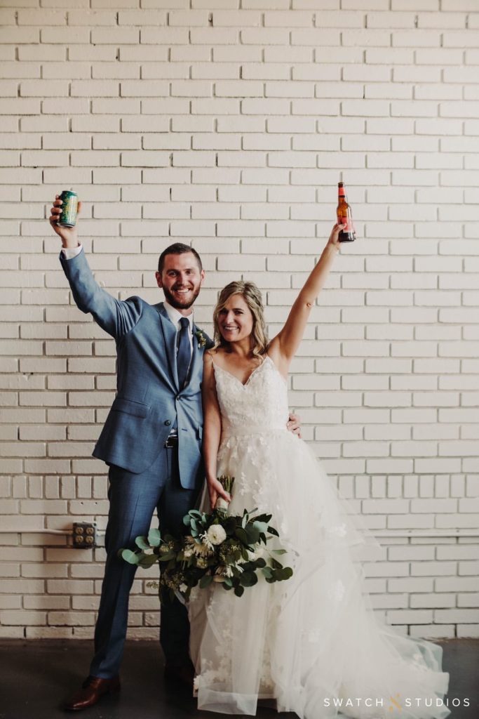 Bride and groom raising glasses to toast in front of a white brick wall at The Venues in Downtown Toledo