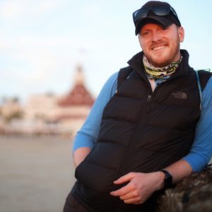 Brennan Stanford leaning against a rock wall wearing a vest and smiling.
