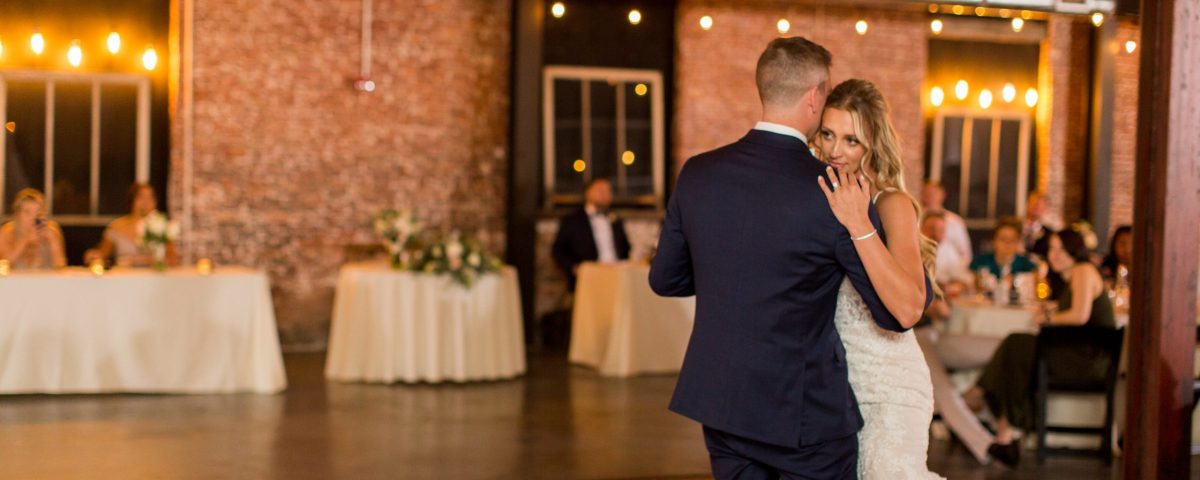 Bride and groom sharing their first dance under bistro lights with a brick backdrop at The Venues Toledo special event venue